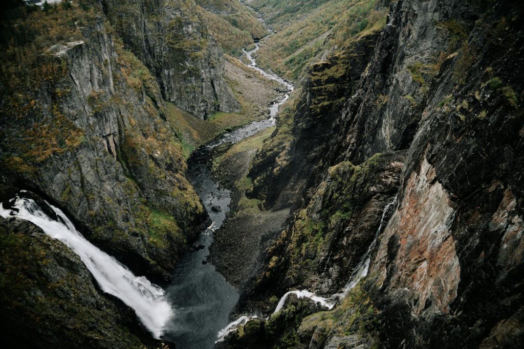 High angle picturesque view of rocky mountains covered with moss located near calm river with waterfall streaming in highland valley