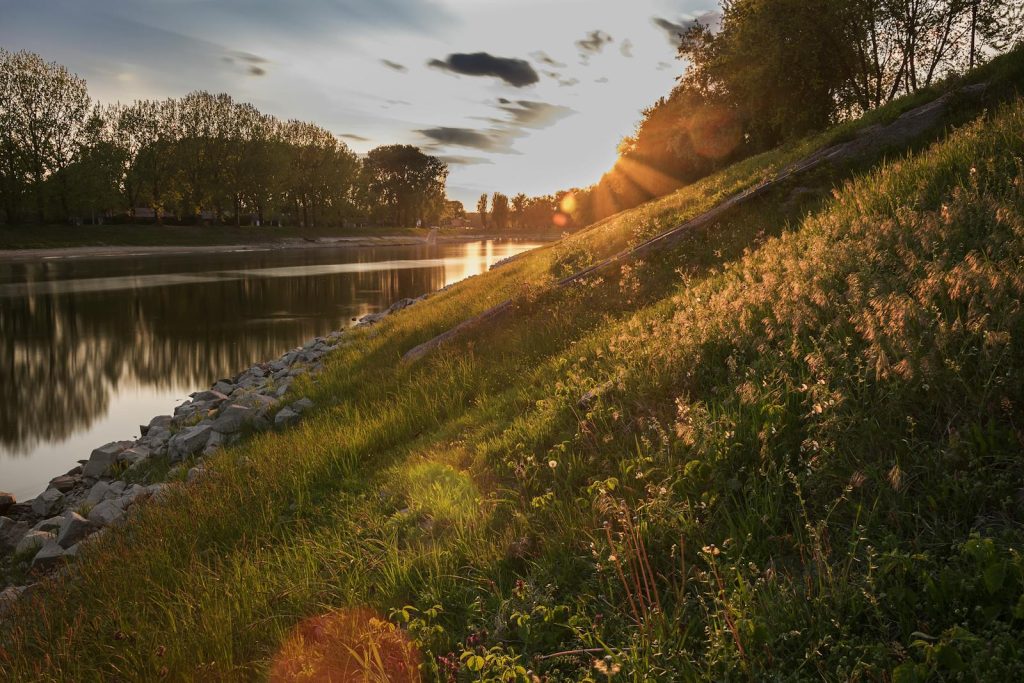 A peaceful sunset scene by a river, with sunlight illuminating the grassy slope and trees in the distance.