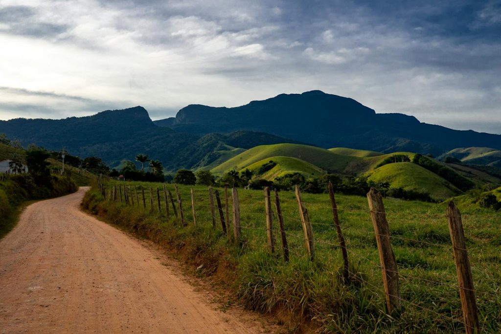 Capture of a dirt road through green hills leading to a mountain range under a cloudy sky.