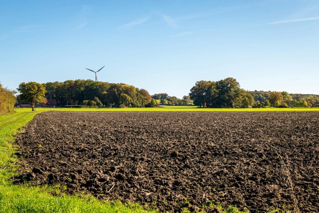 A wind turbine stands near a plowed agricultural field with trees and blue sky in the background.