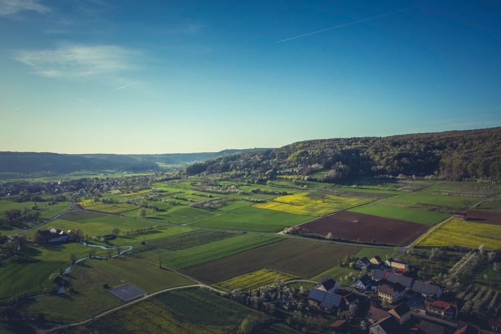 Aerial view of lush green farm fields with a distant village and clear blue sky.